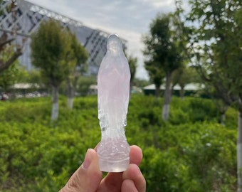 May include: A small, translucent, pink-hued statue of the Virgin Mary. The statue is made of a smooth, polished material and is held in a person's hand.