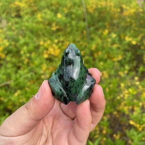 Puede incluir: Una figurita de rana tallada en verde y negro, hecha de piedra preciosa pulida. La rana está sentada con las patas metidas debajo.