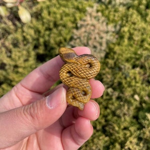 May include: A carved, coiled snake figurine in shades of brown and gold. The snake's scales are intricately detailed, and the head is slightly raised. The figurine is made of a stone-like material and is held in a hand against a blurred green background.