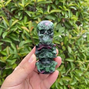 May include: A green and black carved skull and skeleton figurine made of green aventurine gemstone. The figurine has a pink heart on the forehead.