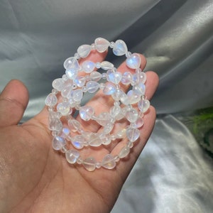 May include: Several strands of heart-shaped, iridescent moonstone beads. The beads are strung together to form bracelets. The moonstone beads have a translucent white color with flashes of blue and rainbow hues. The bracelets are held in a hand.