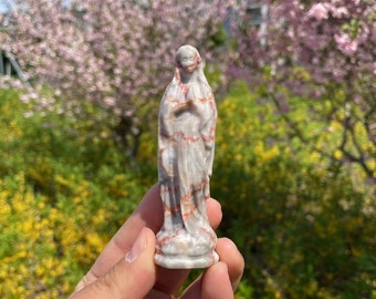 Könnte beinhalten: Eine kleine weiße Marmorstatue der Jungfrau Maria mit roten und braunen Adern. Die Statue wird in der Hand einer Person gehalten.