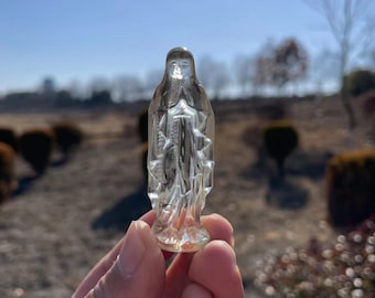 May include: A clear glass figurine of a woman wearing a long robe. The figurine is held in a person's hand.