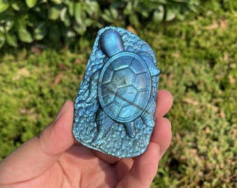 May include: A blue labradorite gemstone carved into the shape of a turtle. The turtle is detailed with a shell, head, and legs. The gemstone has a iridescent sheen.