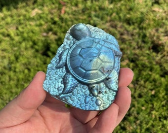 May include: A blue labradorite crystal carved into the shape of a turtle. The crystal has a iridescent sheen and is set on a rough, natural stone base.