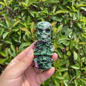 May include: Green and pink gemstone skull carving. The skull is carved with details and has a rough, natural texture.