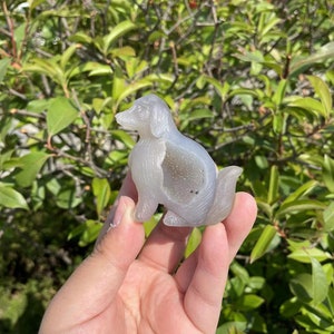 May include: A gray agate carving of a dog. The carving is detailed and has a natural, polished finish. The dog is sitting with its head turned to the side.