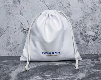 May include: A white drawstring bag made of fabric with two off-white cords. The front of the bag features the blue text KORASY and the smaller brown text Cotten Dust Bags printed in the center.