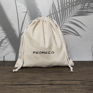 以下が含まれることがあります： 前面に暗い色でかすれたフォントの「PEONECO」という文字がプリントされた、クリーム色の布製巾着袋。上部は2本の白い紐で絞られており、両側に垂れ下がった丸みを帯びた形状をしています。