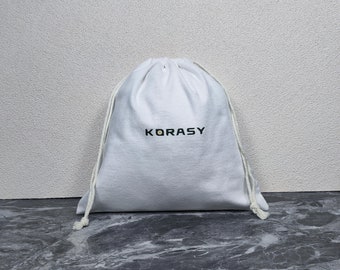 May include: White drawstring bag with the word "KORASY" embroidered in black on the front.