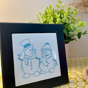 May include: A black framed tile with a white background features a blue illustration of two snowmen. The snowman on the left is wearing a hat and scarf. The snowman on the right is wearing a hat and scarf.