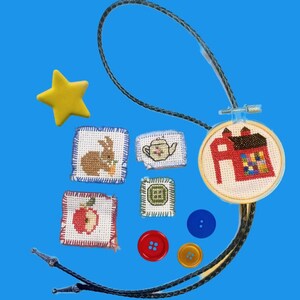 Puede incluir: Una corbata bolo hecha a mano con un cordón trenzado negro y puntas plateadas. La corbata presenta un pequeño aro de bordado de punto de cruz con un diseño de granero rojo. Parches de punto de cruz adicionales incluyen un conejo, una tetera, una manzana y un cuadrado verde. Una estrella amarilla y botones coloridos completan el diseño.