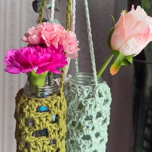May include: Two glass jars with crocheted covers hanging from the ceiling. The jars are filled with pink and purple carnations and a single pink rose. The crocheted covers are green and light green.