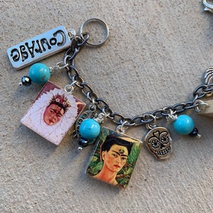 May include: A silver charm bracelet with multiple charms, including a turquoise bead, a silver skull charm, and two square charms with images of Frida Kahlo. The bracelet also features a silver charm with the word "Courage" on it.
