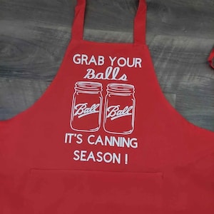 May include: Red kitchen apron with white text that reads "Grab Your Balls It's Canning Season!" and features two Ball mason jars.