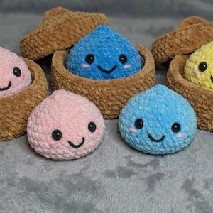 May include: A collection of handmade crochet plushies in various colors, including pink, blue, and yellow. Each plushie has a smiling face and is nestled in a brown basket. The plushies are on a gray surface.