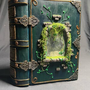 Può includere: Un libro artigianale con copertina e dorso verde scuro. Il libro presenta una finestra decorativa con muschio e viti finte, e chiusure e angoli in metallo ornati. I bordi sono rifiniti in oro.