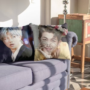 May include: Two decorative throw pillows on a blue sofa. One pillow features a person with silver hair in a black suit. The other pillow has a person wearing glasses and a flower crown. The pillows add a decorative touch to the living room.