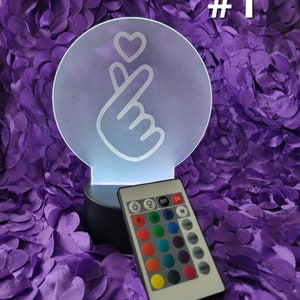 May include: A round, illuminated LED lamp with a hand gesture forming a heart symbol. The lamp has a black base and includes a remote control with color options. The background is a textured purple fabric, and the number "#1" is visible.