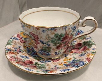 May include: A vintage teacup and saucer set with a floral pattern in shades of pink, red, blue, green, and yellow. The set has a gold trim around the edges.