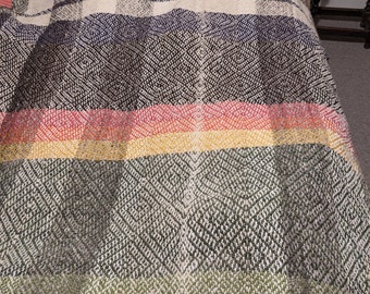 May include: A woven blanket with a colorful striped pattern. The blanket features a mix of colors including green, yellow, red, and black. The blanket is made of a textured material and is draped over a bed.