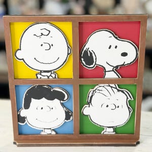 May include: A wooden framed artwork featuring four panels with Peanuts characters. Charlie Brown is on a yellow background, Snoopy on red, Lucy on blue, and Linus on green. The characters are in black and white.