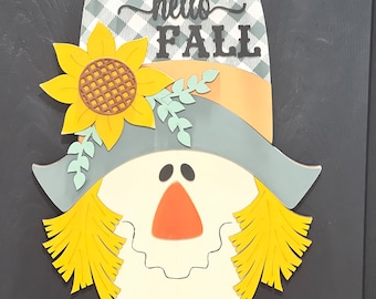 May include: A scarecrow door hanger with a yellow sunflower, a gray hat, and a plaid bow tie. The scarecrow's face is white with a red nose and black eyes. The sign says "hello fall" in black letters.