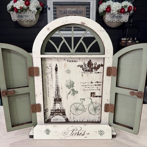May include: A decorative wooden window frame with two hinged shutters in a distressed white and sage green finish. The central panel features a printed graphic of the Eiffel Tower, a bicycle, and the word Paris, accented with copper-toned hinges.
