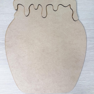 May include: A flat, light brown wooden cutout shaped like a honey pot. The object features a smooth, matte surface with a distinct, wavy drip pattern along the top edge, mimicking the appearance of honey overflowing from the rim of a container.