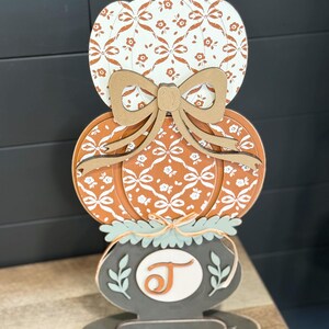 May include: A decorative wooden pumpkin stack with a bow. The top pumpkin is white with a brown floral pattern, the middle pumpkin is orange with a white floral pattern, and the bottom has a gray base with the letter "J".