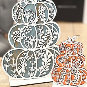 May include: Two wooden pumpkin decorations with intricate cut-out designs. The larger pumpkin is blue and white, while the smaller pumpkin is orange and white.