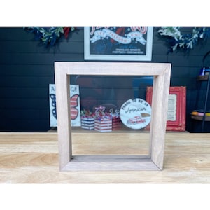 May include: A light wood picture frame with a clear glass insert. The frame is sitting on a wooden table. The frame is empty, but there are red, white, and blue decorations visible through the glass.