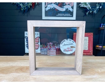May include: A light wood picture frame with a clear glass insert. The frame is sitting on a wooden table. The frame is empty, but there are red, white, and blue decorations visible through the glass.