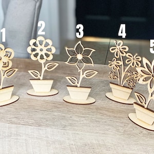 May include: Five wooden flower pot decorations with different flower designs. The flowers are cut out of wood and are attached to a small wooden base. The pots are also made of wood and have a simple design.