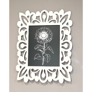 May include: White decorative frame with a cutout design. The frame holds a black and white print of a sunflower.