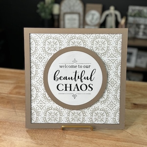 May include: A framed sign with a brown background and a white pattern. The sign has a round cutout with a white background and brown border. The text inside the cutout reads "welcome to our beautiful CHAOS".