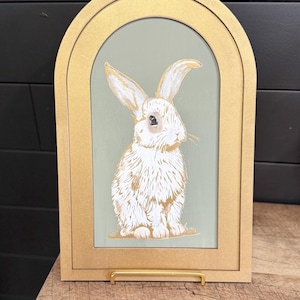 May include: A white rabbit with long ears is painted on a green background. The painting is framed in a gold arch-shaped frame.