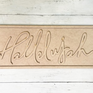 May include: A rectangular wooden sign with the word "Hallelujah" cut out in a cursive font. The sign is made of light brown wood and is set against a light-colored wooden background.