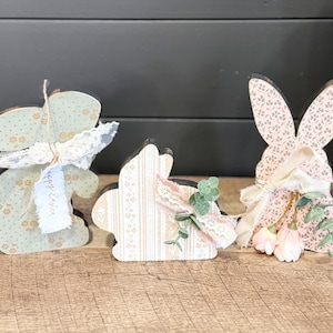 May include: Three wooden Easter bunny decorations. The bunnies are decorated with floral patterned paper in pastel colors. Each bunny has a ribbon and lace embellishment. One bunny has a tag that says "Happy Easter".