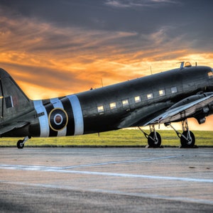 May include: A black and white military aircraft with a roundel on the side, taxiing on a runway at sunset. The aircraft has a white stripe running along the fuselage and wings.