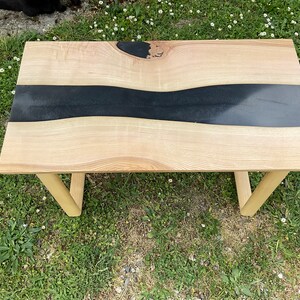 Op de afbeelding: Een houten salontafel met een zwarte epoxyhars rivier die door het midden stroomt. De tafel heeft een natuurlijke houtkleur en eenvoudige houten poten.