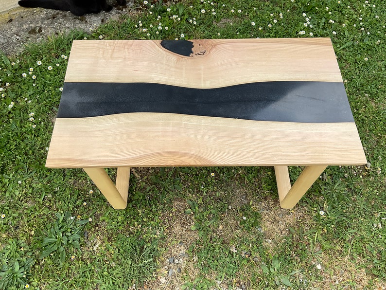 Op de afbeelding: Een houten salontafel met een zwarte epoxyhars rivier die door het midden stroomt. De tafel heeft een natuurlijke houtkleur en eenvoudige houten poten.