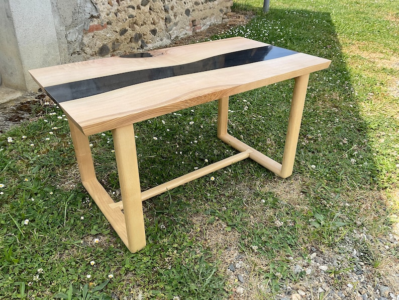 Op de afbeelding: Een houten tafel met een zwarte epoxyharsrivier die door het midden loopt. De tafel heeft een eenvoudig, rechthoekig ontwerp met een lichte houtkleur.