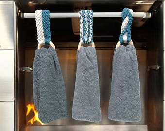 Pode incluir: Três toalhas de cozinha cinzas penduradas em uma barra de metal com laços de crochê branco e turquesa. As toalhas estão penduradas sobre um forno de aço inoxidável com fogo queimando dentro.