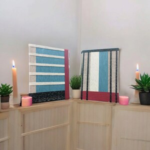 May include: Two wall hangings with a striped pattern in shades of blue, white, and red. The hangings are made of woven yarn and are displayed on a white wall with a wooden shelf.