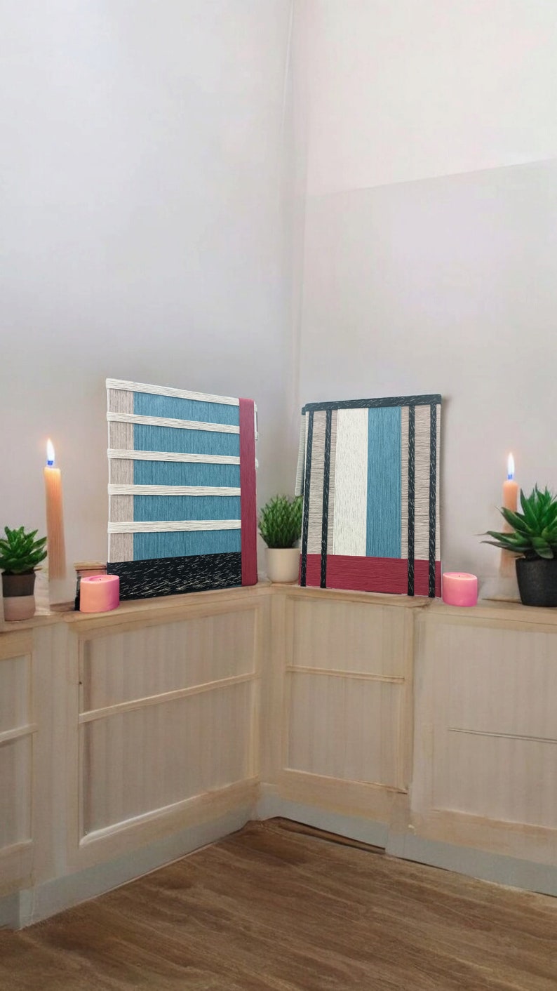 May include: Two wall hangings with a striped pattern in shades of blue, white, and red. The hangings are made of woven yarn and are displayed on a white wall with a wooden shelf.