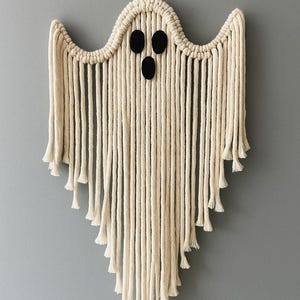 Könnte beinhalten: Ein cremefarbenes Makramee-Geist-Wandbehang. Der Geist hat drei schwarze, ovale Augen und einen gefransten Saum. Aufgehängt an einer naturbelassenen Kordel, ideal für Halloween-Dekoration.