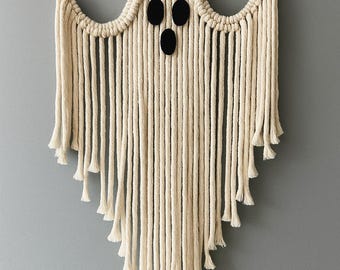 May include: A macrame ghost wall hanging in cream-colored rope. The ghost has three black oval-shaped eyes and a flowing, fringed bottom. The wall hanging is suspended by a natural-colored cord, perfect for Halloween decor.