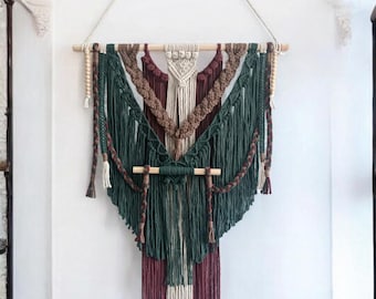 Può includere: Un arazzo da parete in macramè con un design geometrico in tonalità di verde, marrone e bianco. L'arazzo è realizzato con una varietà di nodi e trame, e presenta un'asta di legno nella parte superiore.