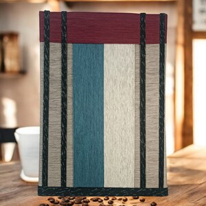 May include: A striped fabric tote bag with a burgundy top, black trim, and a white, blue, and beige striped pattern. The bag is sitting on a wooden table with coffee beans scattered around it.